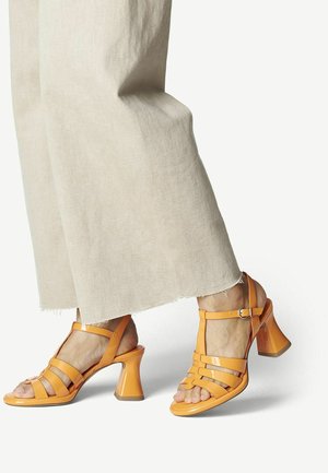 Orange Patentleder-Sandalen mit Blockabsatz, die mehrere Riemen über dem Fuß und einen mit Schnalle versehenen Knöchelriemen aufweisen. Strukturierte Sohle sichtbar.