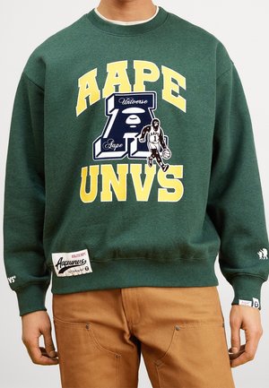 Personne portant un sweat-shirt vert foncé avec un texte jaune "AAPE UNVS" et un graphique d'un joueur de basket-ball sur le devant, assorti à un pantalon marron.