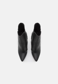 Bottes cuir noir pour femme à la cheville, avec un bout pointu, présentant une texture lisse et une découpe asymétrique. Aucune quincaillerie ni accent visible.