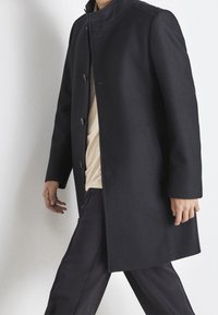 Man loopt, gekleed in een donkerblauwe knopenjas over een beige shirt en bijpassende donkerblauwe broek tegen een lichte achtergrond.