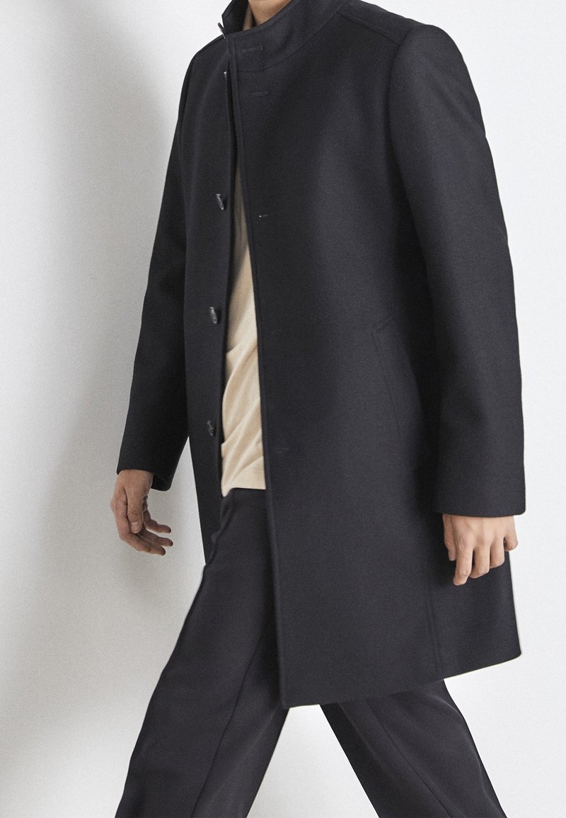 Man loopt, gekleed in een donkerblauwe knopenjas over een beige shirt en bijpassende donkerblauwe broek tegen een lichte achtergrond.