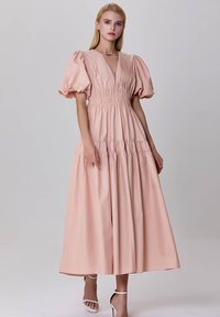 Robe maxi en coton rose avec un corsage smocké, décolleté en V profond et manches bouffantes. Comprend une jupe à volants et des sandales blanches minimalistes.