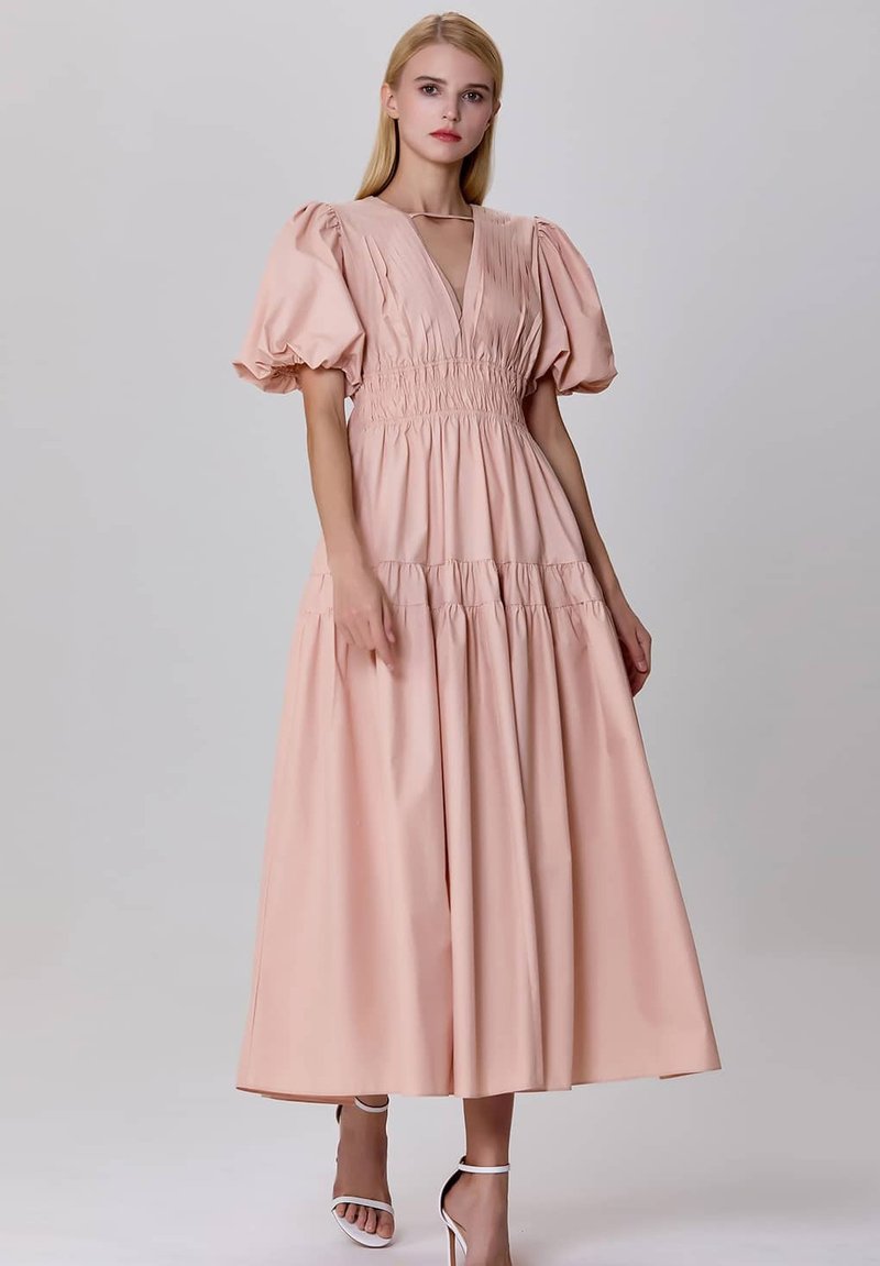 Robe maxi en coton rose avec un corsage smocké, décolleté en V profond et manches bouffantes. Comprend une jupe à volants et des sandales blanches minimalistes.