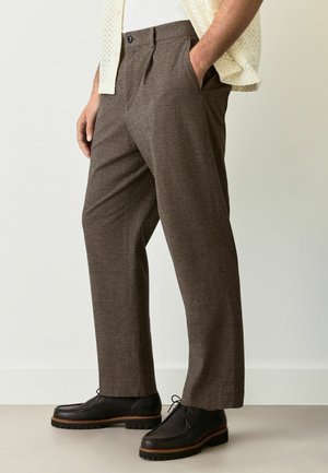 Braune maßgeschneiderte Hose aus strukturiertem Stoff, mit geradem Bein, zwei seitlichen Taschen und einem einzelnen Knopfverschluss. Kombiniert mit schwarzen Schuhen.