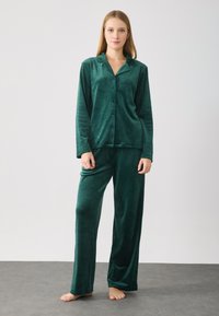 Ungdommelig kvinne står barføtt på grått gulv iført pyjamas-sett i mørkegrønn velour med lange ermer og bukser.