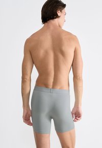 Šedé boxerky s hladkou texturou a elastickým pasem, zobrazené zobrazené zezadu na modelu na jednoduchém bílém pozadí.