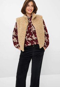 Beige nepbont vest met hoge kraag over een gestreepte blouse in rood en crèmewit, gecombineerd met zwarte corduroy broek. Getextureerd en gelaagd ontwerp.
