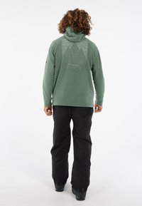 Groene fleecetruien met hoge kraag, met een bergafbeelding op de achterkant en een gladde textuur. Gekleed met zwarte skibroeken en laarzen.