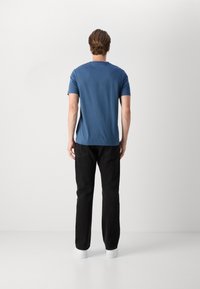 T-shirt blu a maniche corte da uomo con colletto rotondo, abbinato a pantaloni neri a gamba dritta e sneakers bianche a bassa altezza, in piedi su uno sfondo neutro.