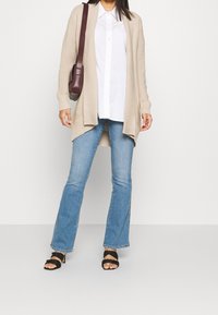 Cardigan beige a coste sopra una camicia bianca con bottoni, abbinato a jeans a zampa leggera blu chiaro e sandali neri con tacco. Borsa a spalla marrone come accessorio.