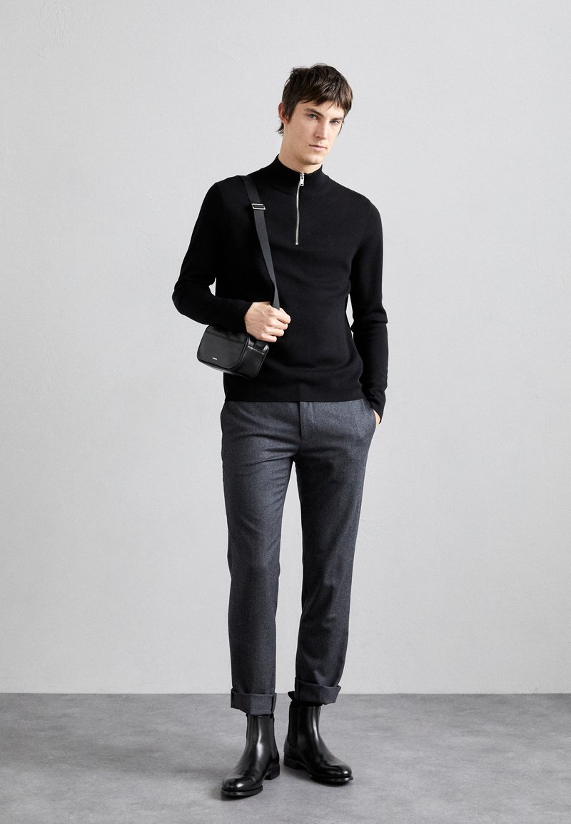 Black zip-neck sweater, gray tailored trousers, and black ankle boots. Carries a small black crossbody bag. Smooth texture, minimal design.