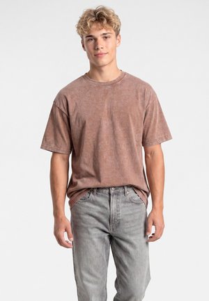 Jonge man met krullend blond haar, draagt een vervaagd bruin T-shirt en lichtgrijze spijkerbroek, staande tegen een effen witte achtergrond.