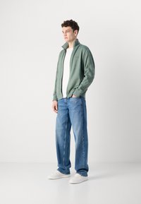 Grønn zip-up jakke med høy krage, i kombinasjon med hvit skjorte og blå jeans. Hvite joggesko fullfører antrekket. Glatt stofftekstur.