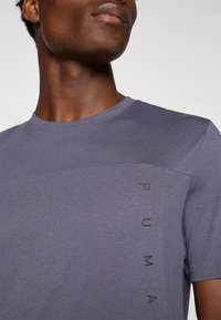 Szara bawełniana koszulka z krótkim rękawem, z delikatną fakturą i pionowo nadrukowanym logo "PUMA" z boku. Okrągły dekolt.