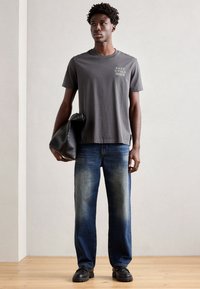 Grijze katoenen T-shirt met ronde hals en witte tekstprint, gecombineerd met vervaagde blauwe rechte jeans en een zwarte leren schoudertas. Zwarte schoenen.