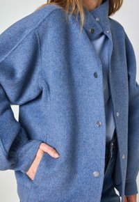 Giacca blu in misto lana con collo, bottoni a pressione e tasche laterali; presenta una texture morbida e una vestibilità ampia.