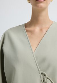 Jasnozielona bluzka o kroju kopertowym, wykonana z gładkiego materiału, z głębokim dekoltem w kształcie litery V, szerokimi rękawami oraz wiązaniem w talii.