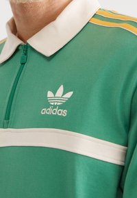 Grön zip-up polo skjorta med vit krage och rand över bröstet. Har den ikoniska vita Adidas-logotypen och gula axelränder.