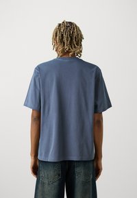 Tricou din bumbac de culoare albastră, cu guler rotund și mâneci scurte. Prezintă o croială lejeră, o textură netedă și detalii minime de cusătură.