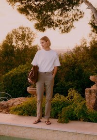 Femme debout près d'une piscine au coucher du soleil, portant un t-shirt blanc, un pantalon beige, des tongs, tenant une veste marron, avec des arbres et des arbustes derrière elle.