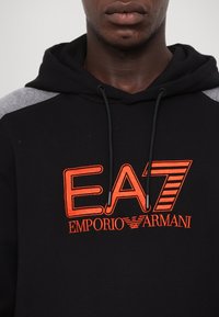 Felpa nera con dettagli grigi. Presenta un logo ricamato arancione "EA7 EMPORIO ARMANI" nella parte anteriore. Coulisse per il cappuccio regolabile.
