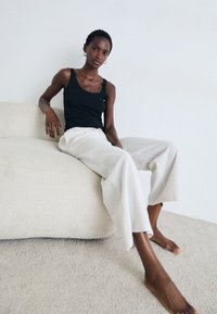 En smal person som bär en svart linne och lösa krämfärgade byxor sitter barfota på en texturerad vit soffa mot en enfärgad vägg.