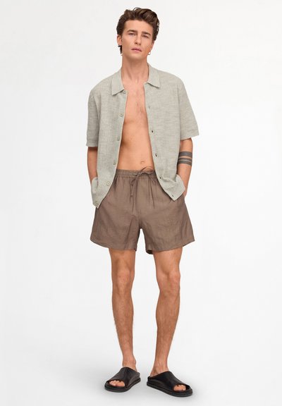 Homme portant une chemise beige à manches courtes ouverte, un short marron à cordon, des sandales noires à enfiler, avec un tatouage sur l’avant-bras gauche, debout sur un fond blanc.