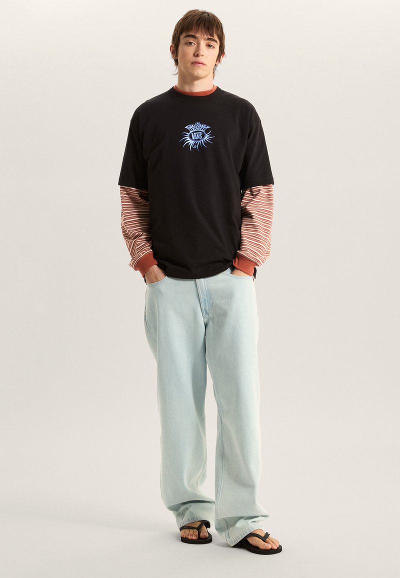 Camiseta negra con gráfico azul, superpuesta sobre una camiseta de manga larga a rayas rojas y blancas. Jeans de pierna ancha azul claro y chanclas negras.