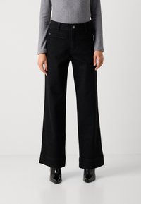 Jeans de pierna ancha negros con una cintura alta, elaborados en denim, con un ligero brillo, bolsillos frontales y dobladillo enrollado. Combinados con botas de tobillo brillantes.