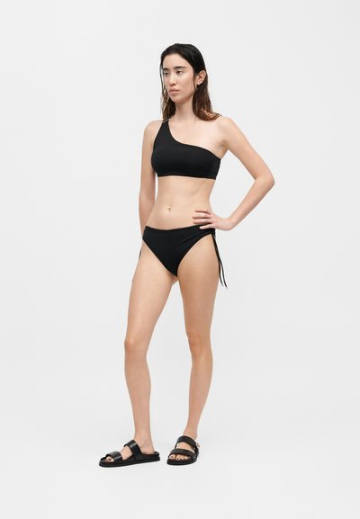 Γυναίκα με μαύρο μπικίνι με έναν ώμο και ταίριασμα κάτω μέρος με κορδόνια στο πλάι, στέκεται με το χέρι στη μέση και μαύρα σανδάλια σε απλό φόντο.