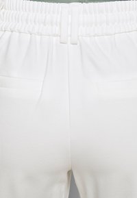 Bílé kalhoty s elastickým pasem a hladkou texturou, které mají dvě boční kapsy a uvolněný střih. Úhledné prošívání podél švů.