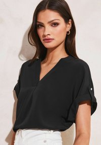 Blusa nera a maniche corte con scollatura a V, realizzata in tessuto morbido, e con risvolti alle maniche che presentano bottoni a pressione. Il modello indossa pantaloni bianchi a vita alta.