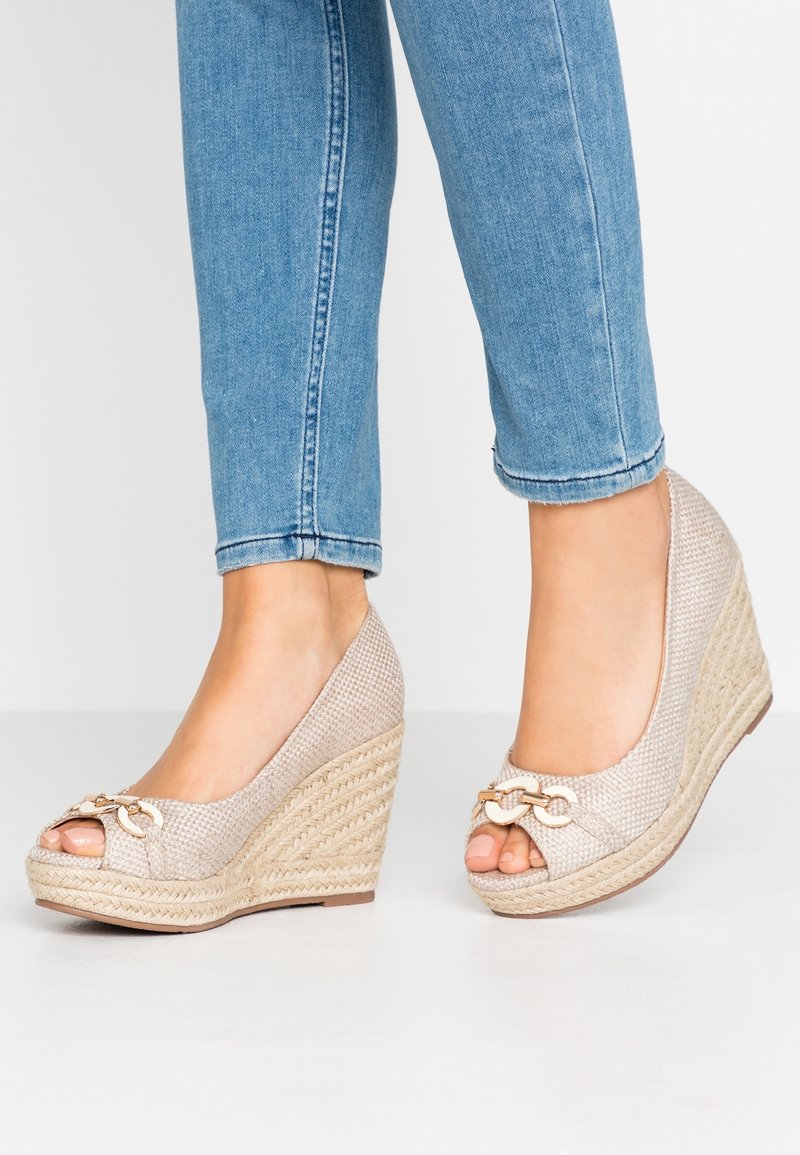 Pieds portant des talons compensés beige tissés avec un détail de chaîne dorée et orteils ouverts, associés à un jean skinny bleu clair retroussé.