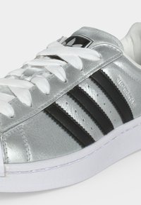 Ασημένιο αθλητικό παπούτσι Adidas Superstar με μαύρες ρίγες, λευκά κορδόνια και λευκή λαστιχένια σόλα, εμφανιζόμενο σε κοντινή πλευρική άποψη.