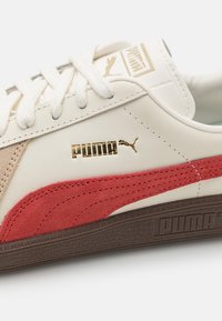 Tenisky Puma s bílým koženým svrškem a červenými a béžovými semišovými akcenty. Obsahují zlaté logo, ploché bílé tkaničky a texturovanou hnědou podrážku.