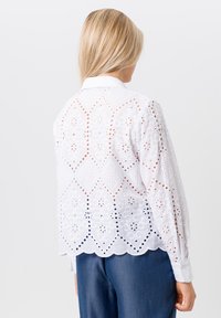 Witte, geborduurde blouse met uitgesneden bloemmotieven, lange mouwen en een collerneck. Golvende zoomafwerking.
