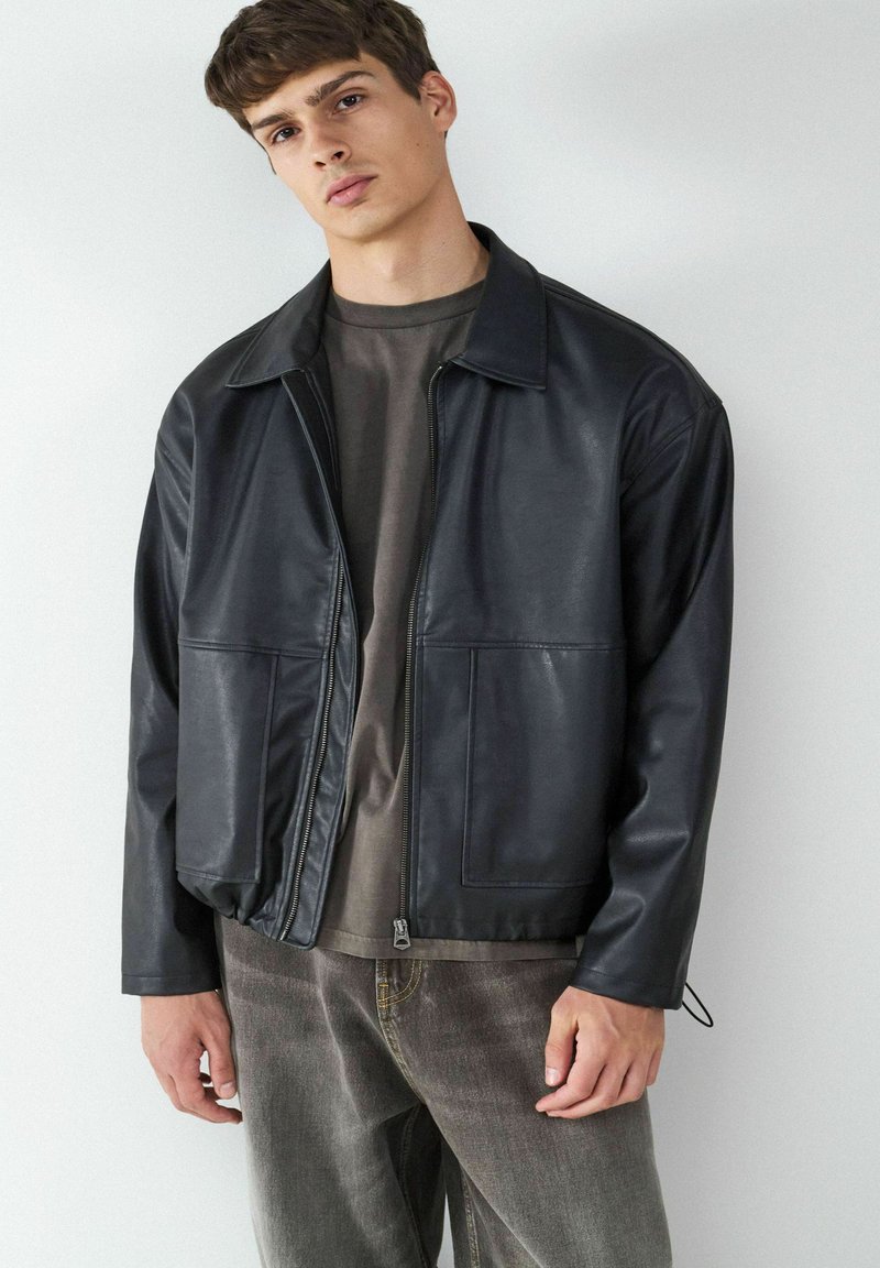 Giubbotto in pelle nera con collo a camicia, chiusura con zip, due tasche anteriori e vestibilità comoda, indossato sopra una maglietta grigia scura e jeans grigi.