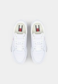 Fehér sneaker perforált felsőrésszel, lapos fűzőkkel és piros, fehér és kék színű logóval. Simább textúra és kerek orrforma.