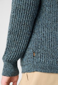 Strickpullover in Dunkelblau und Petrol mit gerippter Textur. Verfügt über ein kleines oranges "BOSS"-Etikett an der linken Hüfte.