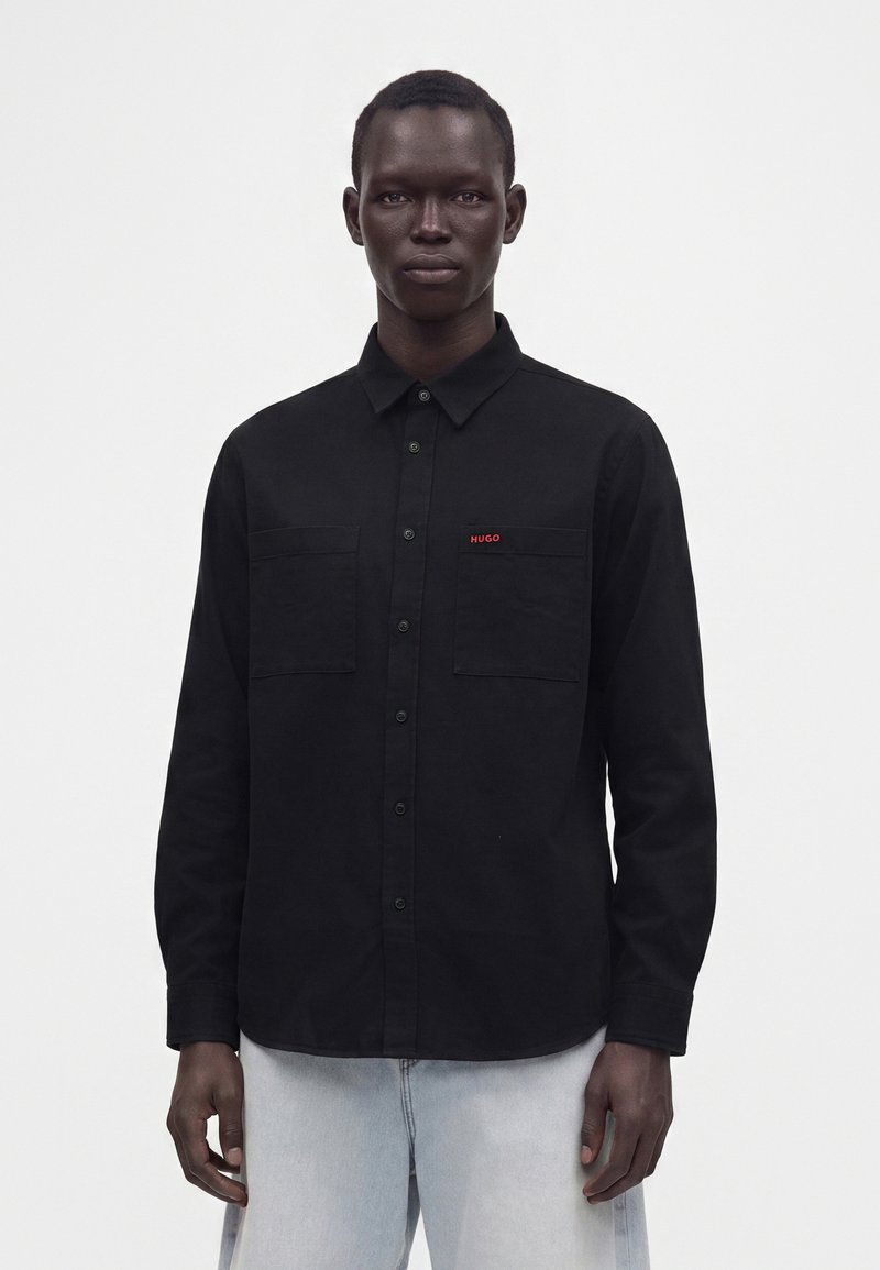Homme portant une chemise noire à boutons avec deux poches avant, logo rouge "HUGO" sur la poitrine gauche, et un jean bleu clair sur fond blanc.