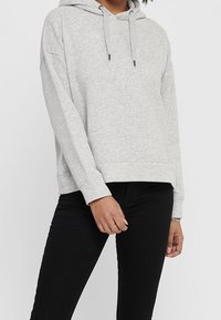 Felpa con cappuccio grigio chiaro realizzata in tessuto morbido. Presenta una tasca frontale a marsupio e lacci sul cappuccio. Indossata con jeans neri.