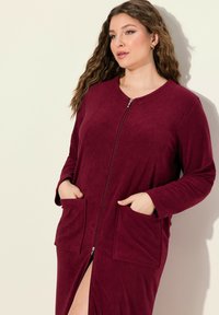 Accappatoio bordeaux con zip realizzato in tessuto morbido, con maniche lunghe, due tasche frontali e uno spacco laterale fino alla coscia.
