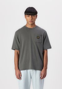 Gray cotton t-shirt with a round neck and short sleeves, featuring a visible left chest pocket and yellow branding detail.