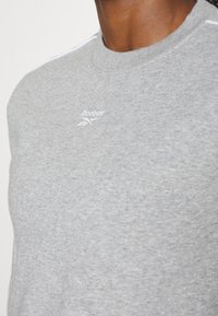 Gros plan sur une personne portant un sweat-shirt Reebok gris clair avec un petit logo blanc Reebok sur la poitrine.