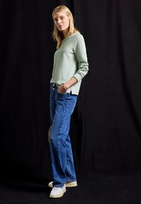 Jasnozielona bluza z teksturowanym wykończeniem, z bocznymi rozcięciami, połączona z niebieskimi dżinsami o prostym kroju i białymi sneakerami na czarnym tle.