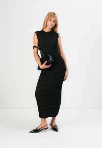 Vrouw in een gestructureerde mouwloze zwarte midi-jurk, met een zwarte handtas, draagt zwarte slingback kitten heels en staat tegen een witte achtergrond.