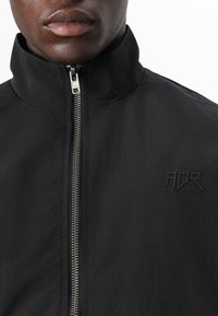 Schwarze Zip-Jacke mit hohem Kragen und strukturiertem Stoff. Verfügt über einen silbernen Reißverschluss und ein dezentes schwarzes "ADR"-Logo auf der Brust.