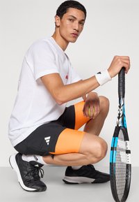 Fehér póló, fekete és narancssárga sportnadrág, fekete sportcipő. Teniszkötelet és labdát tart, ülve egy összpontosító arckifejezéssel.