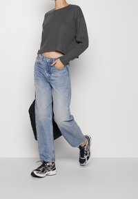 Maglione corto grigio scuro, jeans larghi di media tonalità e sneakers nere con dettagli argentati. Lo sfondo è semplice e neutro.