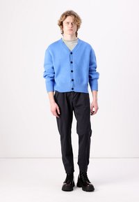 Cardigan albastru deschis cu un tricot texturat, cu decolteu în V, mâneci lungi și nasturi închis la culoare, purtat cu pantaloni negri tapered și cizme.